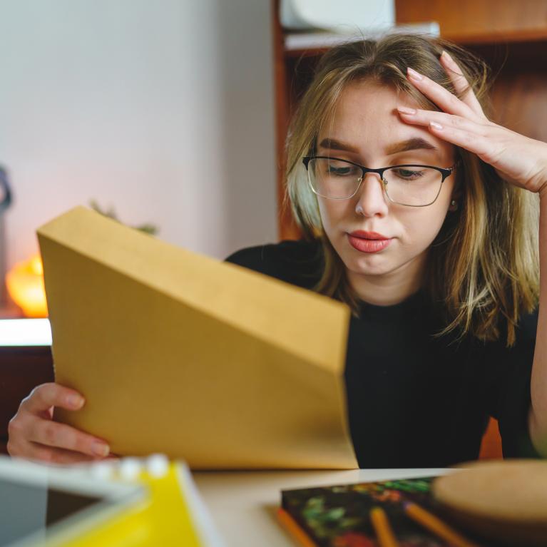 Jente eller ung kvinne med blondt og hår og briller som ser stresset ut og holder papir i den ene hånda