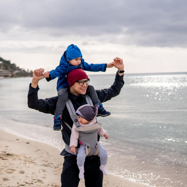Pappa på tur på stranden med ett barn i bæresele og ett på nakken