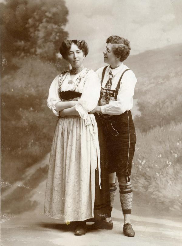 Two women in a black and white photo, one is dressed in traditonal women's clothes, the other in men's clothes Two women holding around each other in a black and white photo, one is dressed in traditonal women's clothes, the other in men's clothes